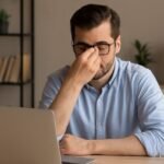 clean wooden desk with an open laptop in a minimalist home office setting, featuring a bookshelf with books and a small plant in the background, and a lamp on a side table.